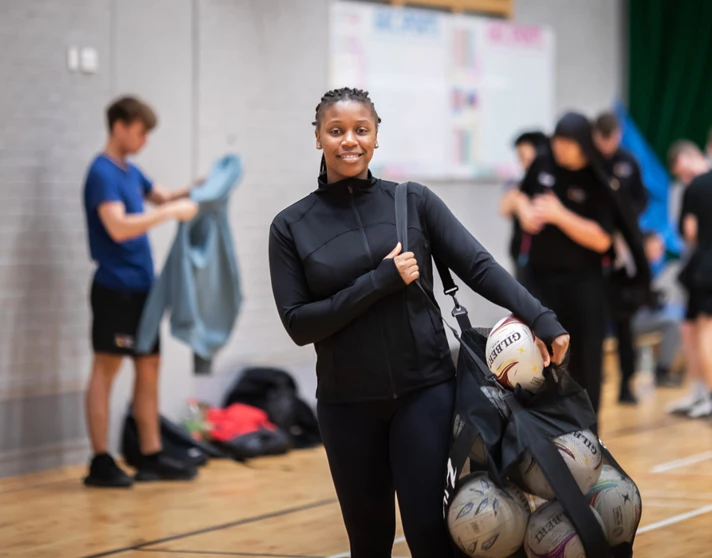 A student holding a mesh bag filled with netballs, smiling at the camera in a sports hall. A student holding a mesh bag filled with netballs, smiling at the camera in a sports hall.